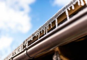 Hanging icicles from the roof of a wooden building on a winter frosty day, a lot of snow on the roof, a visible plastic gutter and blue sky.
